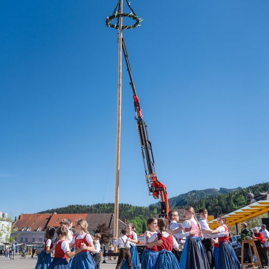 Eine Gruppe von Menschen in traditionellen Kostümen tanzt um einen Pfahl mit einem Kran, der einen Kranz in der Mitte eines Stadtplatzes hebt.