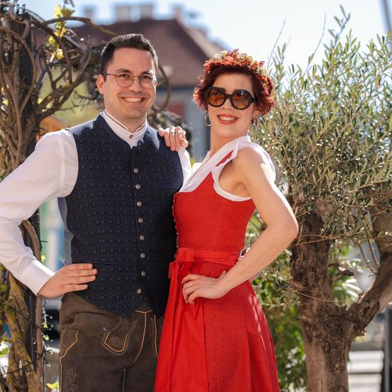 Ein Mann und eine Frau stehen lächelnd vor einem Baum. Er trägt eine Brille und eine Weste, sie trägt ein rotes Kleid und eine Sonnenbrille.