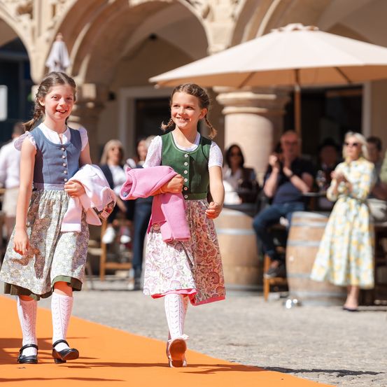 Zwei junge Mädchen gehen gemeinsam in einem traditionellen Festumfeld, lächelnd und halten rosa Stoff. Dahinter beobachtet eine Menge unter einem Regenschirm.