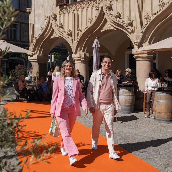 Ein Paar in aufeinander abgestimmten rosa Anzügen geht auf einem orangefarbenen Teppich vor einem historischen Gebäude, mit sitzenden und stehenden Personen in der Nähe.