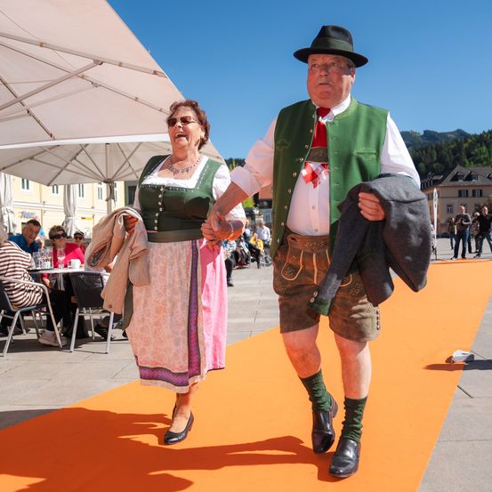 Ein älteres Paar geht einen Pfad hinunter, beide in traditioneller bayerischer Kleidung. Der Mann trägt eine grüne Weste und braune Shorts, während die Frau ein Blumenkleid trägt.