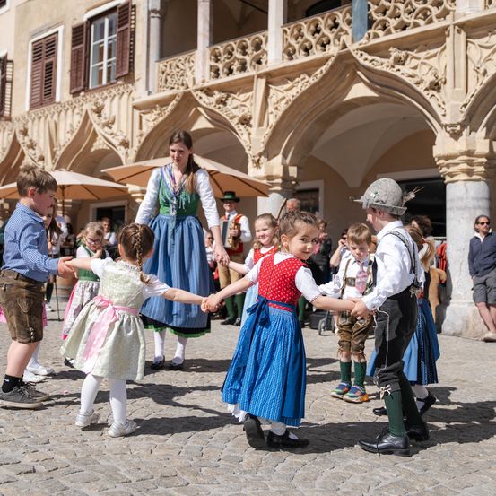 Kinder in traditioneller Kleidung tanzen im Kreis in einer Außenumgebung mit einem historischen Gebäude im Hintergrund.