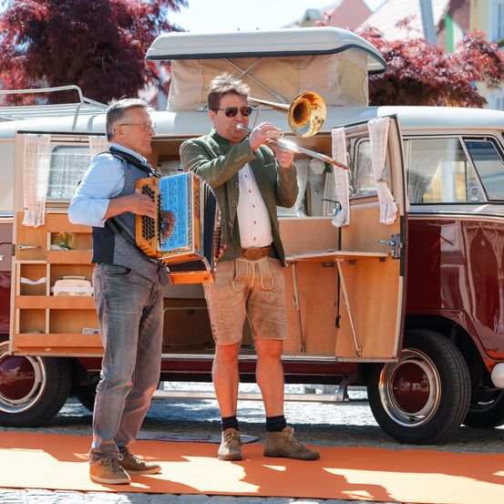 Zwei Männer spielen Musik vor einem Oldtimer-Van. Einer spielt Posaune, der andere Akkordeon. Beide tragen lässige Kleidung. Der Van hat offene Türen, die sein Inneres zeigen.