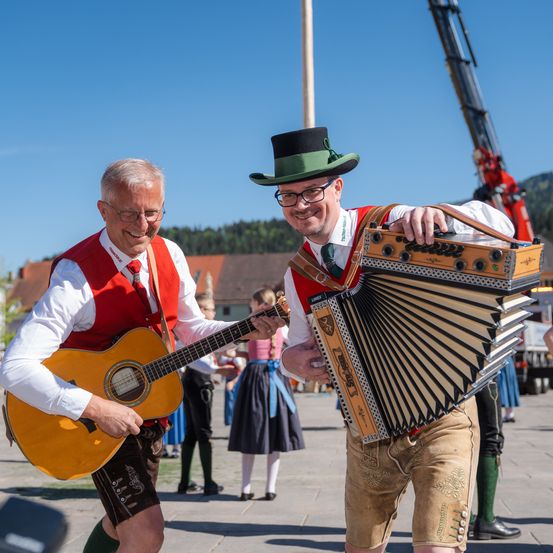 Zwei Männer spielen traditionelle Instrumente auf einem Stadtplatz, einer spielt Gitarre und der andere Akkordeon, beide in traditioneller Kleidung, mit einem Kran im Hintergrund.