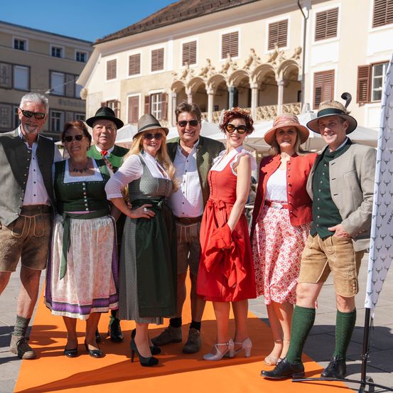 Eine Gruppe von Menschen, die in traditioneller bayerischer Kleidung gekleidet sind, stehen gemeinsam auf einem roten Teppich vor einem historischen Gebäude. Sie lächeln für ein Foto.