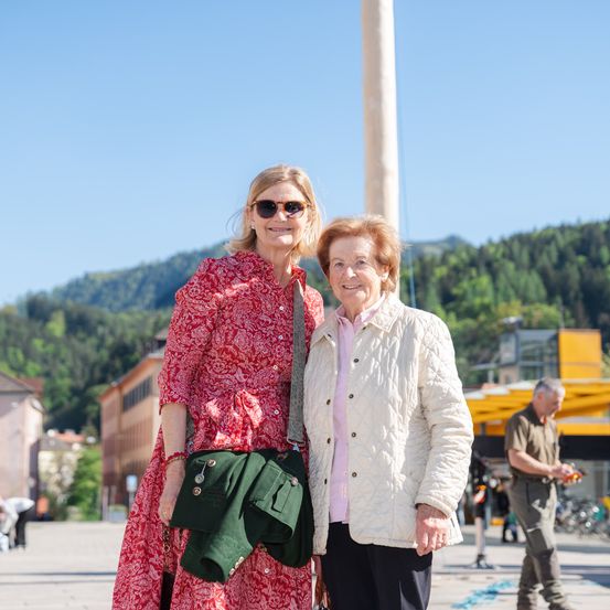 Zwei Frauen stehen lächelnd auf einer Straße. Eine trägt ein rotes gemustertes Kleid und eine grüne Tasche. Hinter ihnen ist ein Pfahl und Gebäude zu sehen. In der Ferne Berge und ein weiterer Mann mit einem Fahrrad.