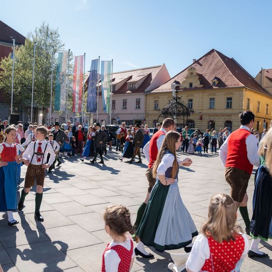 Eine Gruppe von Menschen in traditioneller deutscher Kleidung tanzt mitten auf der Straße. Im Hintergrund Gebäude mit Flaggen.