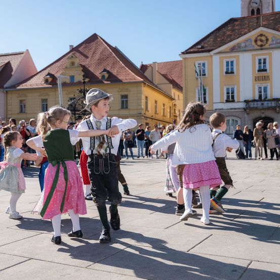 Eine Gruppe von Kindern tanzt mitten auf einem Platz vor Gebäuden. Einige tragen traditionelle Kleidung.