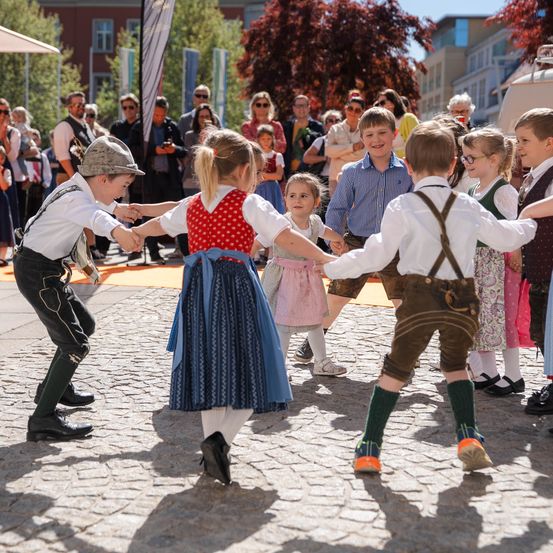 Kinder in traditionellen Kostümen tanzen im Kreis auf einer Kopfsteinpflasterstraße, während ein Publikum zuschaut.