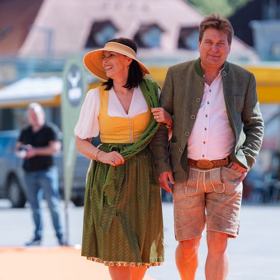 Ein Mann und eine Frau in traditioneller deutscher Kleidung gehen zusammen auf einer Straße. Hinter ihnen macht ein Mann Fotos.