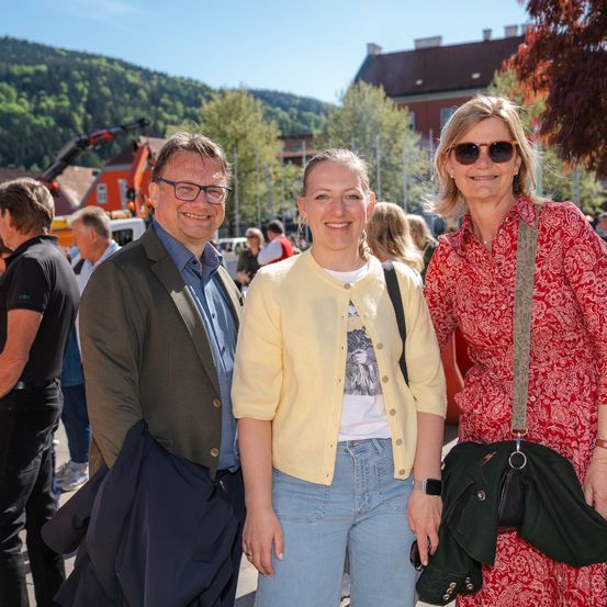 Drei Erwachsene stehen vor einer malerischen Bergkulisse im Freien. Zwei Frauen tragen Sonnenbrillen und eine trägt eine Tasche. Ein Mann hält eine Jacke.