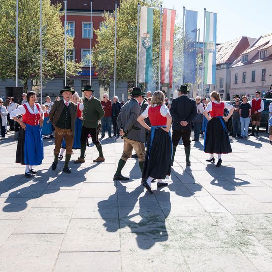 Eine Gruppe von Menschen in traditioneller Kleidung tanzt auf einem Platz mit Gebäuden und Flaggen im Hintergrund.