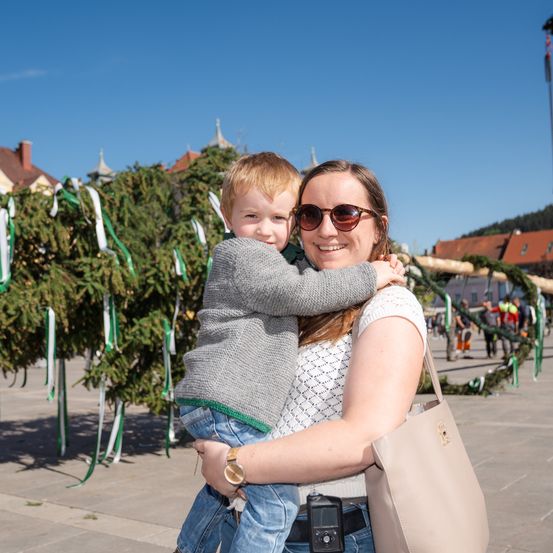 Eine Frau hält einen lächelnden Jungen vor einem geschmückten Baum, beide tragen eine Sonnenbrille. Sie stehen draußen in der Nähe eines Betonwegs mit Grünpflanzen im Hintergrund.