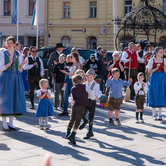 Eine Gruppe von Menschen, einschließlich Kinder und Erwachsene, tanzt in einem Freiluftbereich, wahrscheinlich bei einem Festival. Sie sind in traditioneller Kleidung gekleidet.