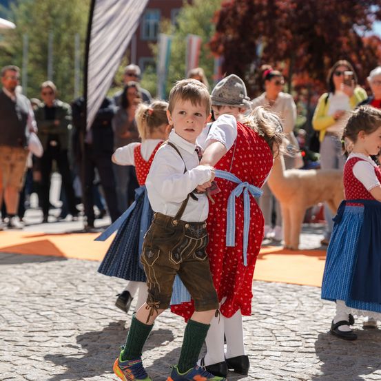 Eine Gruppe von Kindern in traditioneller deutscher Kleidung tanzt auf einer Kopfsteinpflasterstraße. Ein Junge in grünen Socken führt ein Mädchen in einem rot gepunkteten Kleid. Dahinter schauen Menschen zu.