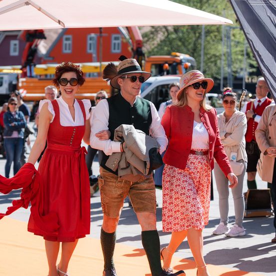 Ein Mann und zwei Frauen in traditioneller Kleidung gehen in einer Außenumgebung, mehrere Personen und Fahrzeuge im Hintergrund.