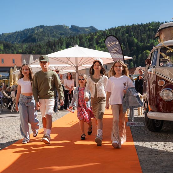 Eine Gruppe von Menschen, einschließlich Kindern, geht auf einem orangefarbenen Teppich draußen. Einer hat einen Hut, und sie sind in der Nähe eines Vans und von Sonnenschirmen. Es gibt sitzende und stehende Personen in der Nähe.