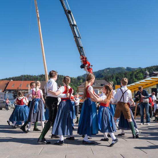 Eine Gruppe von Menschen in traditionellen Kostümen tanzt im Kreis, mit einem Kran und Bergen im Hintergrund.