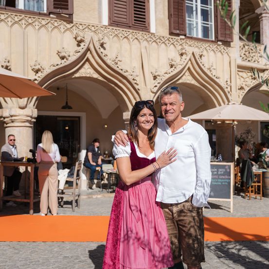 Ein Paar steht vor einem traditionellen Gebäude und lächelt für ein Foto. Hinter ihnen sitzen und stehen Menschen in der Nähe von Tischen und Stühlen. Das Gebäude verfügt über Bogenfenster und -türen.