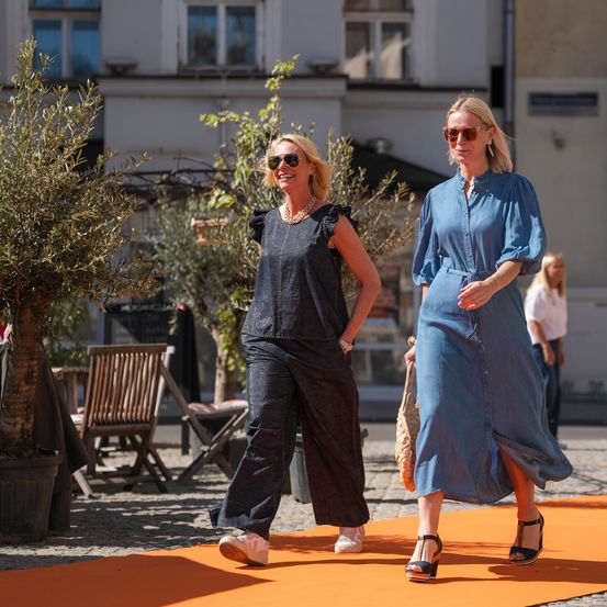 Zwei Frauen, beide lächelnd, gehen vor einem Gebäude auf der Straße. Eine trägt ein dunkles Jumpsuit, die andere ein blaues Kleid. Sie sind von Topfpflanzen und einem orangefarbenen Teppich umgeben.