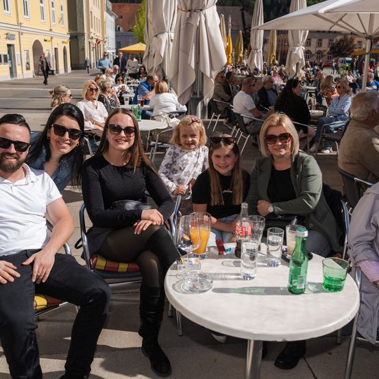Eine Gruppe von Menschen sitzt draußen an einem Tisch mit Getränken und Snacks. Ein junges Mädchen und eine Frau in einer Jacke sitzen in der Nähe. Gebäude und eine Menschenmenge sind im Hintergrund.