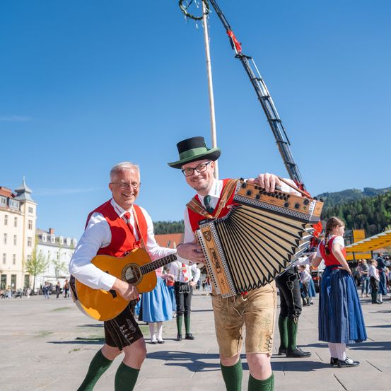 Zwei Männer spielen Gitarre und Akkordeon auf einem Platz. Einer trägt eine rote Weste, grüne Socken und einen Hut. Der andere trägt grüne Shorts und einen grünen Hut. Hinter ihnen sind Menschen, Gebäude und ein Kran.