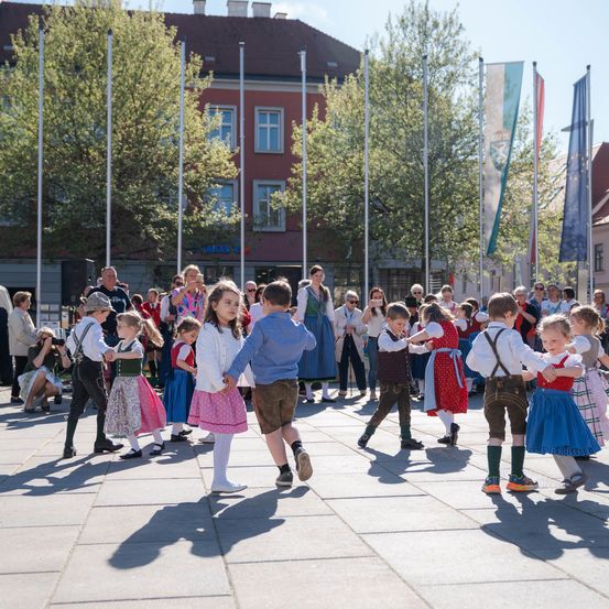 Kinder in traditioneller bayerischer Kleidung tanzen auf einem offenen Platz, umgeben von Zuschauern, Fahnen und Gebäuden.