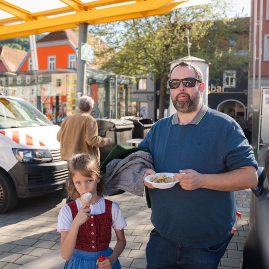 Ein Mann mit einem Teller Essen und ein Mädchen, das Eis isst, stehen vor einem Auto und einer Bushaltestelle.