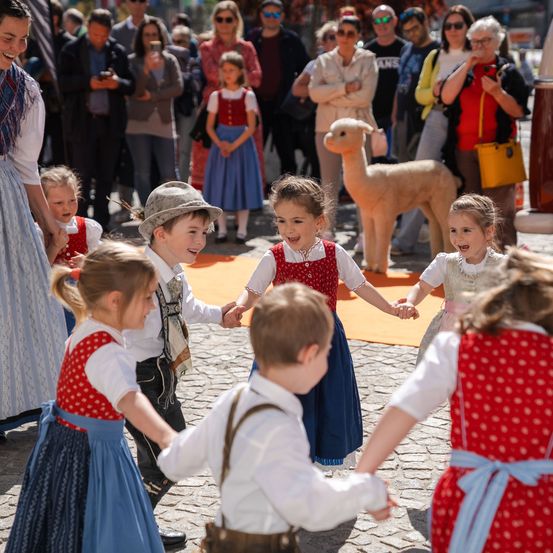 Mehrere Kinder, gekleidet in traditioneller Kleidung, halten Händchen und tanzen im Kreis. Sie sind von einer Menge umgeben, die von hinten zuschaut. Im Hintergrund befindet sich ein pelziges Tier und eine Gruppe von Menschen, von denen einige Fotos machen.