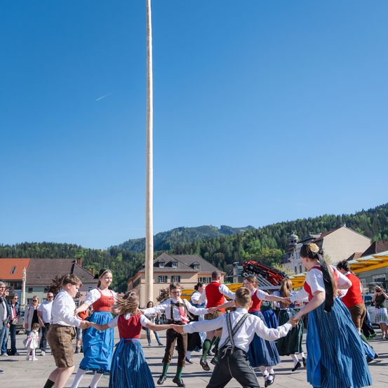 Eine Gruppe von Menschen tanzt auf einem Platz und trägt traditionelle Kleidung. Hinter ihnen steht ein hoher Pfahl mit Gebäuden und Bergen im Hintergrund.