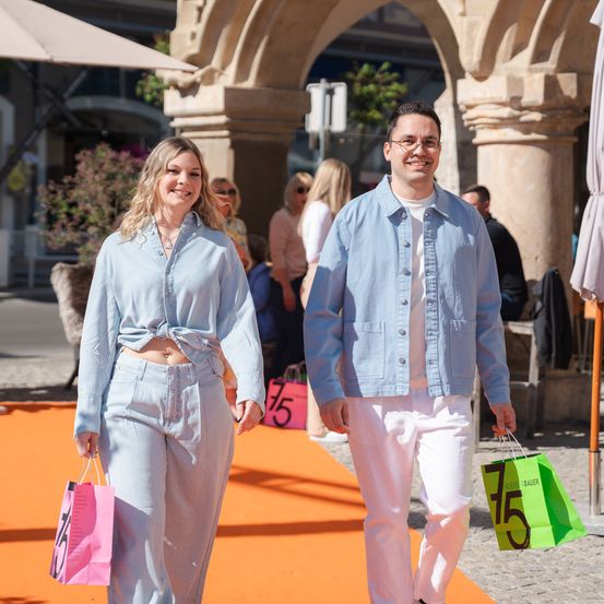 Ein Mann und eine Frau, lächelnd, gehen Hand in Hand auf einem orangefarbenen Teppich und halten Einkaufstüten mit Rabattetiketten. Sie sind in Hellblau gekleidet und stehen vor einem Bogen. Dahinter schlendern und sitzen weitere Menschen.