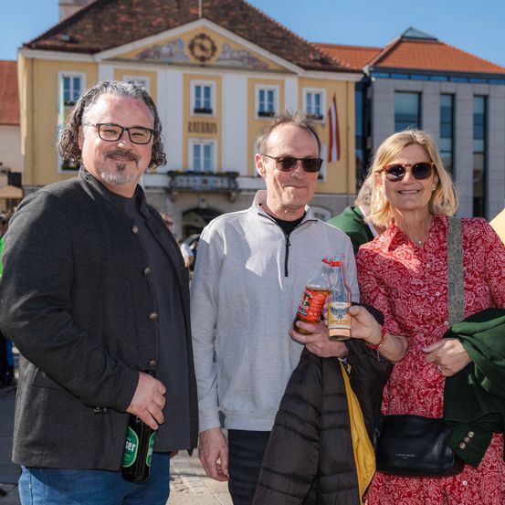 Drei Menschen stehen vor einem Gebäude. Ein Mann und eine Frau. Die Frau hält zwei Flaschen. Sie tragen alle eine Sonnenbrille und scheinen zu lächeln.