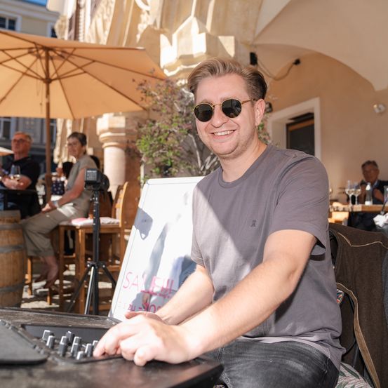 Ein Mann mit Sonnenbrille sitzt in einem Außencafé, lächelt und schaut auf ein elektronisches Gerät auf dem Tisch. Hinter ihm sitzt eine Frau an einem Tisch mit Weingläsern.
