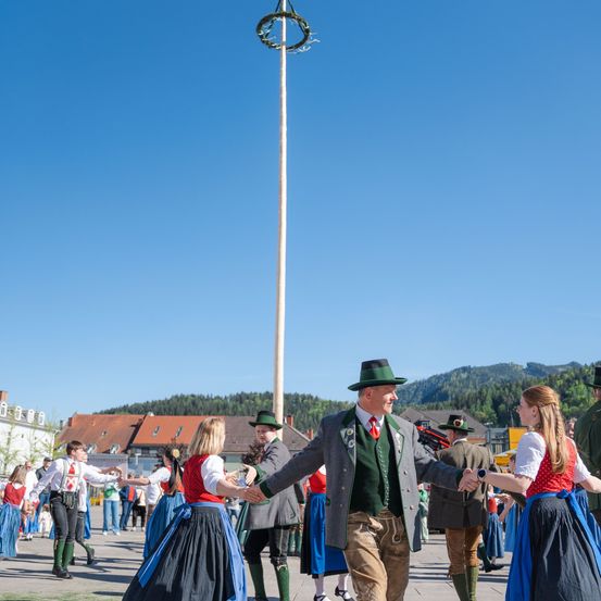 Eine Gruppe von Menschen in traditioneller Kleidung tanzt an einem sonnigen Tag im Kreis. Ein Mann mit grünem Hut führt eine Frau in einem roten Kleid. Ein hoher Pfahl mit einem dekorativen Kranz steht hinter ihnen. Gebäude und Berge sind im Hintergrund.