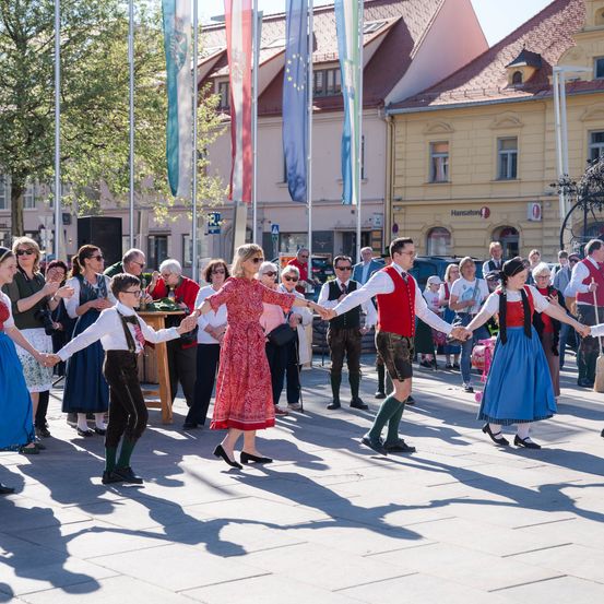 Eine Gruppe von Menschen in traditioneller Kleidung tanzt auf einem Platz. Gebäude mit Flaggen säumen die Straße, und Zuschauer schauen zu. Bäume und geparkte Fahrzeuge ergänzen die Szene.