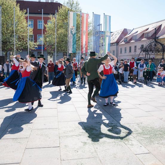 Menschen in traditioneller Kleidung tanzen auf der Straße. Flaggen und Gebäude sind im Hintergrund. Zuschauer beobachten sie an einem sonnigen Tag.