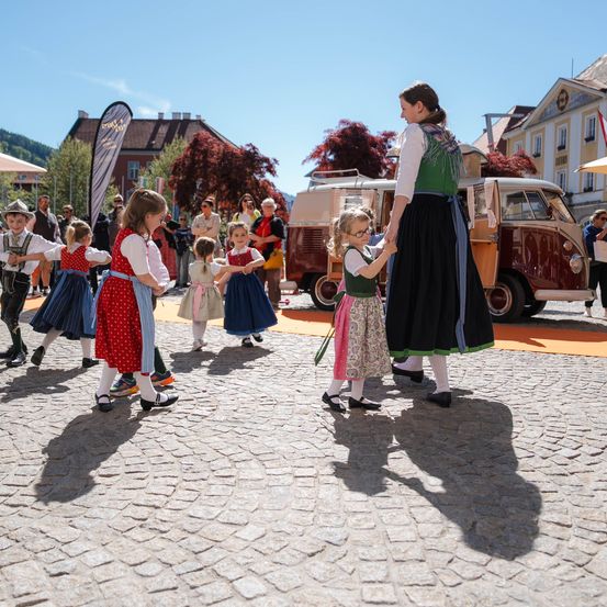 Eine Gruppe von Kindern in traditioneller Kleidung tanzt um eine Frau, die ein kleines Mädchen an der Hand hält, auf einer Kopfsteinpflasterstraße. Dahinter ist ein alter Van geparkt, und mehrere Leute beobachten von den Seitenlinien.