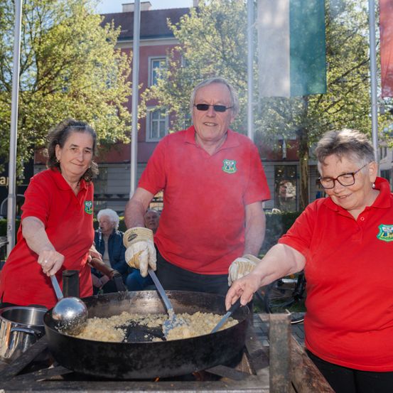 Drei Personen in roten Hemden bereiten ein Essen über einem offenen Feuer im Freien zu. Eine Person rührt, während die anderen zusehen. Ein Gebäude und Bäume sind im Hintergrund.
