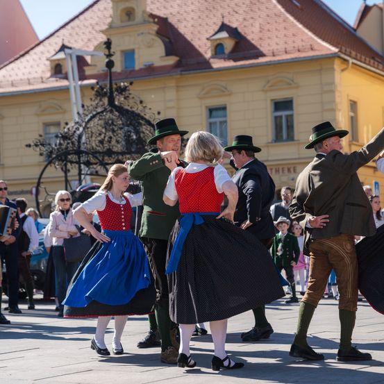 Eine Gruppe von Menschen in traditionellen Kostümen tanzt auf einem Platz vor einem Gebäude.