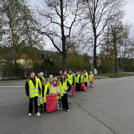 Eine Gruppe von Menschen, einige Kinder, tragen reflektierende Westen und Handschuhe und stehen in einer Reihe. Sie halten rote Taschen. Dahinter stehen Bäume, ein Zaun und ein Gebäude.