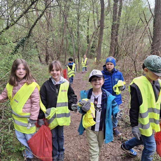 Eine Gruppe von Kindern, die gelbe Westen und Handschuhe tragen, stehen in einem Wald und lächeln für ein Foto. Sie halten Beutel, wahrscheinlich zum Sammeln von Müll. Bäume umgeben sie, mit weiteren Kindern im Hintergrund.