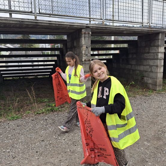 Zwei junge Mädchen in Sicherheitswesten und Handschuhen sammeln Müll auf einem Schotterweg in der Nähe einer Brücke. Sie halten rote Tüten mit Zeichnungen und lächeln.