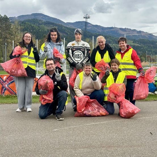 Eine Gruppe von Menschen in Reflektorwesten, die rote Tüten halten, posiert für ein Foto. Sie stehen auf einem gepflasterten Bereich mit einem Grasbereich dahinter. In der Ferne sind Berge zu sehen.