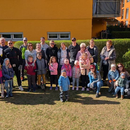 Eine Gruppe von Menschen, einschließlich Erwachsener und Kindern, posiert für ein Foto vor einem gelben Gebäude. Einige tragen Mützen, Brillen und Turnschuhe. Ein Busch steht hinter ihnen.