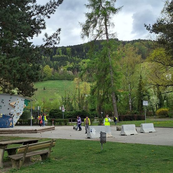 In einem Park gehen Menschen mit einigen Taschen spazieren. In der Nähe befindet sich eine Kletterwand auf der linken Seite und eine Holzbank. Ein Mülleimer und Betonbarrieren liegen auf dem Boden. Bäume und Büsche sind in der Umgebung, und ein Berg ist in der Ferne.