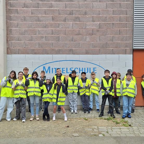 Eine Gruppe von Schülern in gelben Warnwesten posiert für ein Foto vor dem Gebäude der Mielsschule. Sie lächeln und machen Gesten. Die Wand hinter ihnen hat ein blaues Logo.