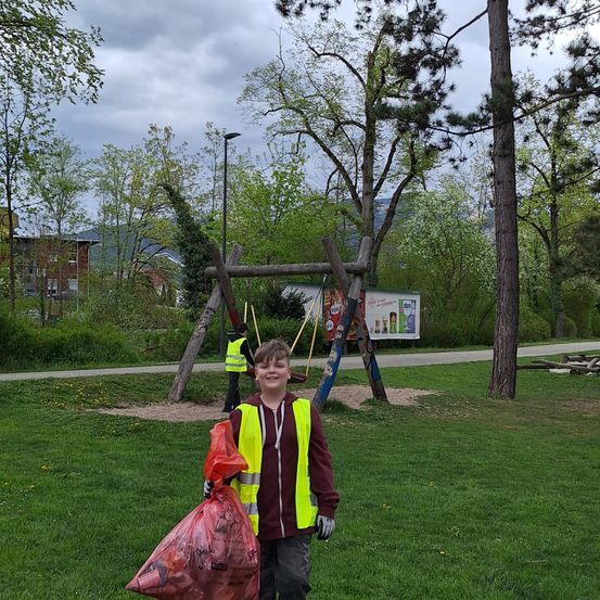 Ein junger Junge in einem gelben Warnweste hält einen roten Beutel in einem Park mit Bäumen und einer Schaukel im Hintergrund. Eine andere Person in einer gelben Warnweste reinigt das Gebiet.