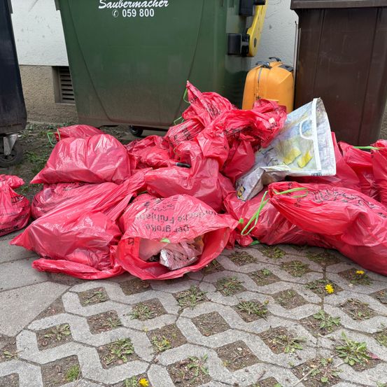 Ein Haufen roter Plastiktüten, einige mit Müll gefüllt, liegt auf einem Ziegelweg neben grünen und braunen Mülltonnen.