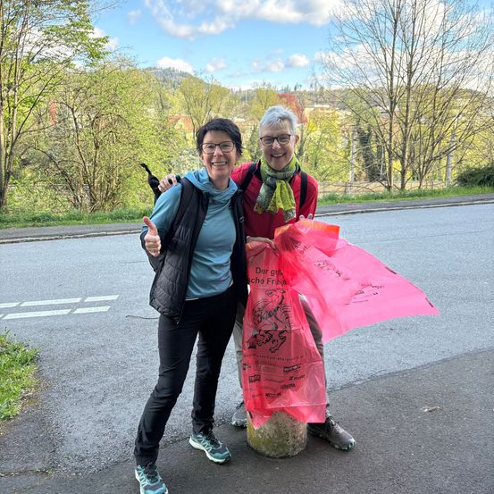 Zwei Frauen in Outdoor-Kleidung stehen auf einer Straße mit Bergen im Hintergrund. Eine hält eine rosa Tasche und lächelt.