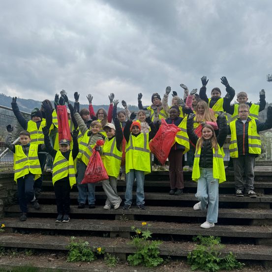 Eine Gruppe von Kindern und Erwachsenen, gekleidet in gelbe Westen, Handschuhe und mit roten Tüten, stehen auf einer Freitreppe im Freien und posieren für ein Foto.